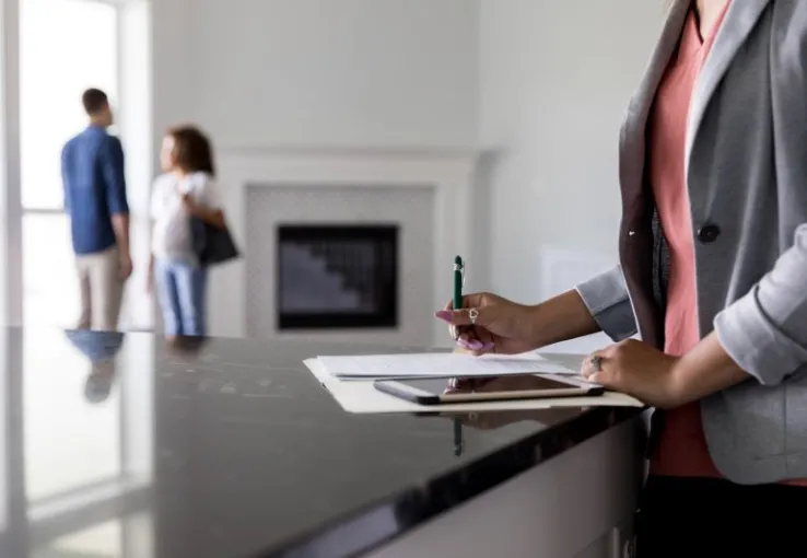 Couple in the background discussing the potential of buying a new house with the agent in the foreground filling out paperwork.